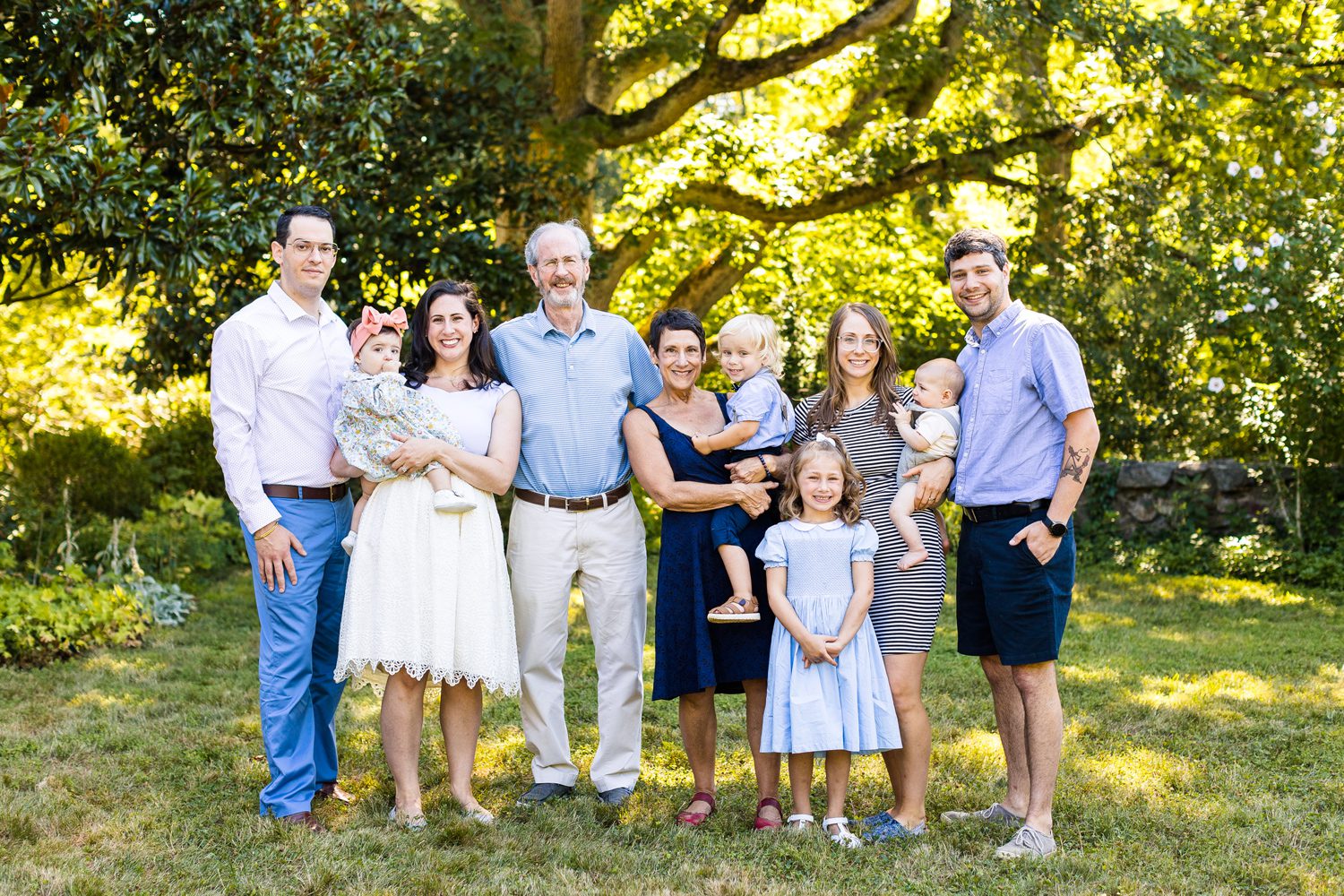 Extended family photo with grandparents, daughters with their husbands, and four grandchildren outside with green trees in background